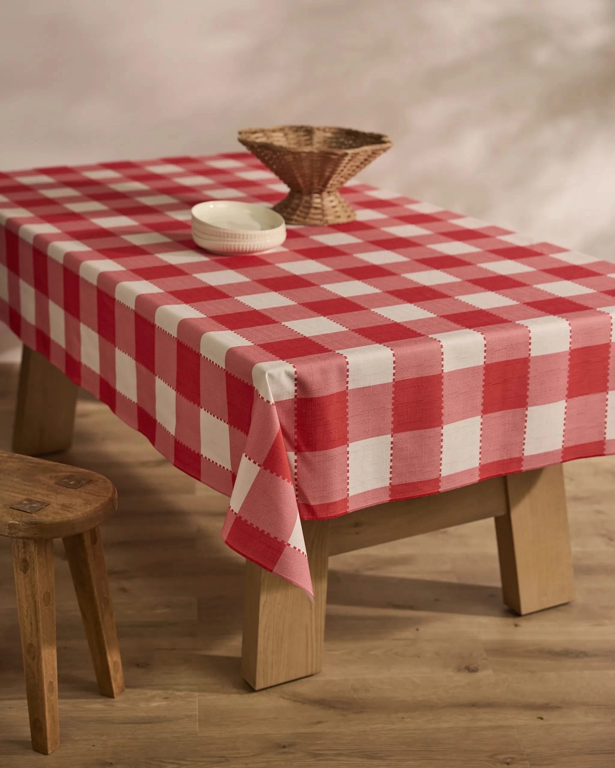 A wooden table is draped with the Geometry Ripe Tomato Gingham stain-resistant tablecloth, set with a small wicker basket and white dish. A wooden stool is beside it, all atop a light wood floor.