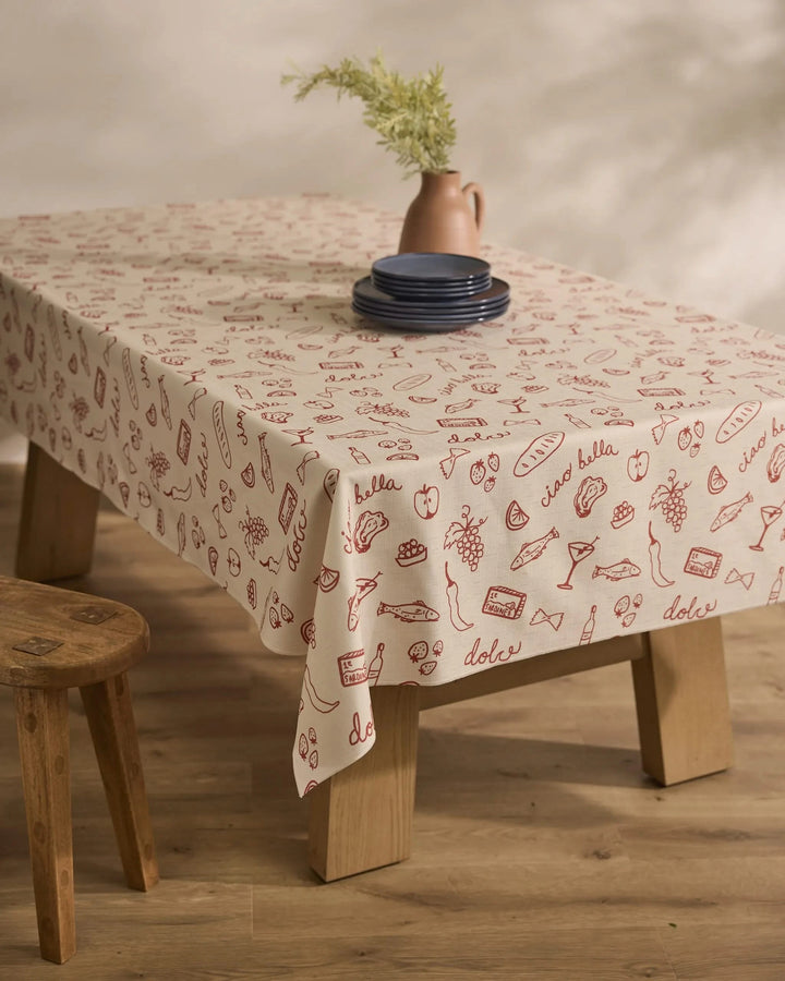 A wooden table is draped with Geometrys Summer Air Tablecloth, featuring red food-themed doodles and Italian words, topped with blue plates and a pink vase of greenery. A wooden stool sits nearby on the wood floor.