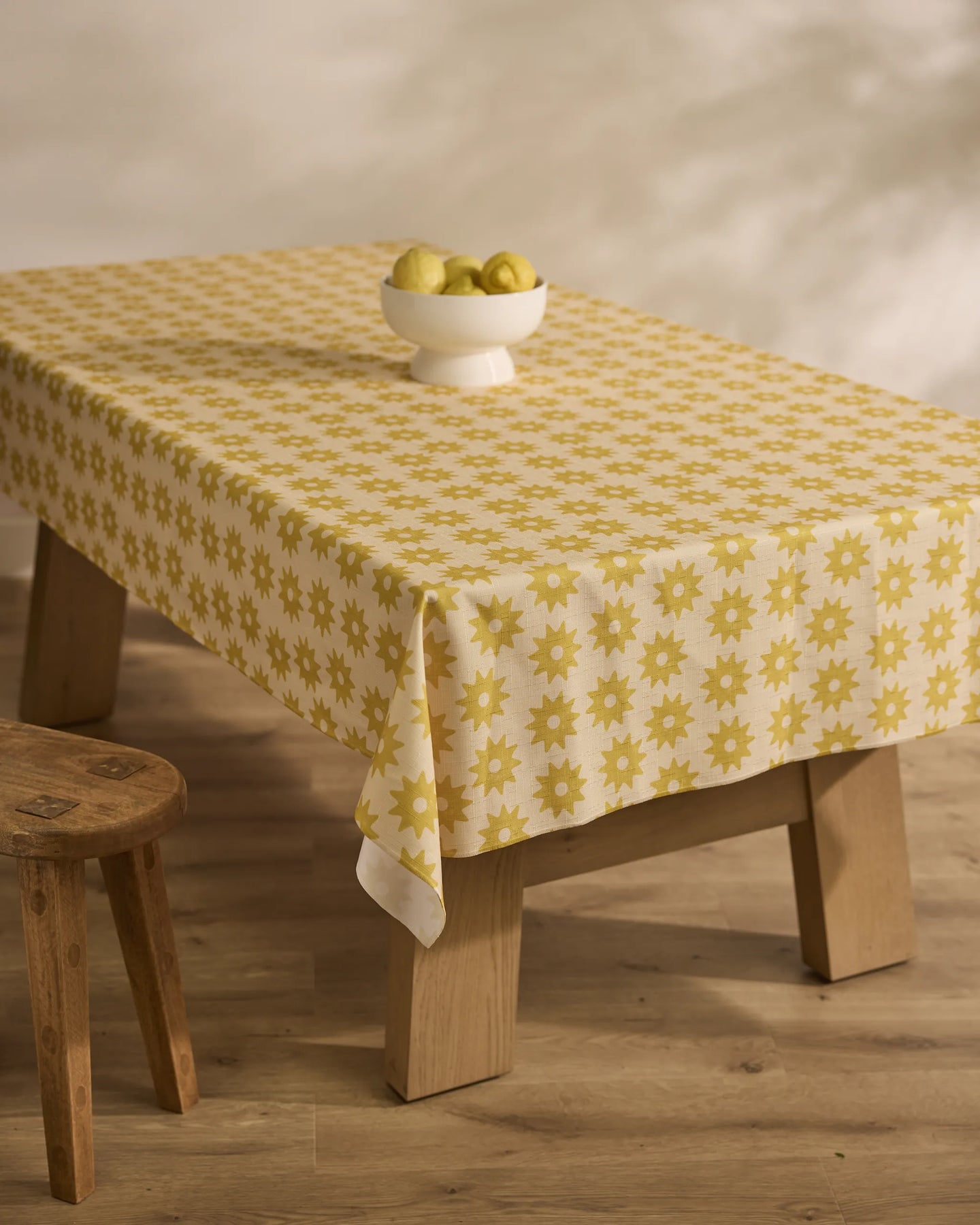 A wooden table is draped with the Geometry Sunny Sun lightweight yellow and white floral-patterned tablecloth. A white bowl of green pears sits on top, next to a wooden stool on a light wood floor.
