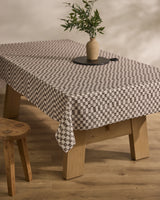 A Tavern wooden table by Geometry is topped with a stain-resistant black-and-white checkered tablecloth, a round black tray, and a ceramic vase with green leafy branches; a wooden stool sits beside it on a light wood floor.