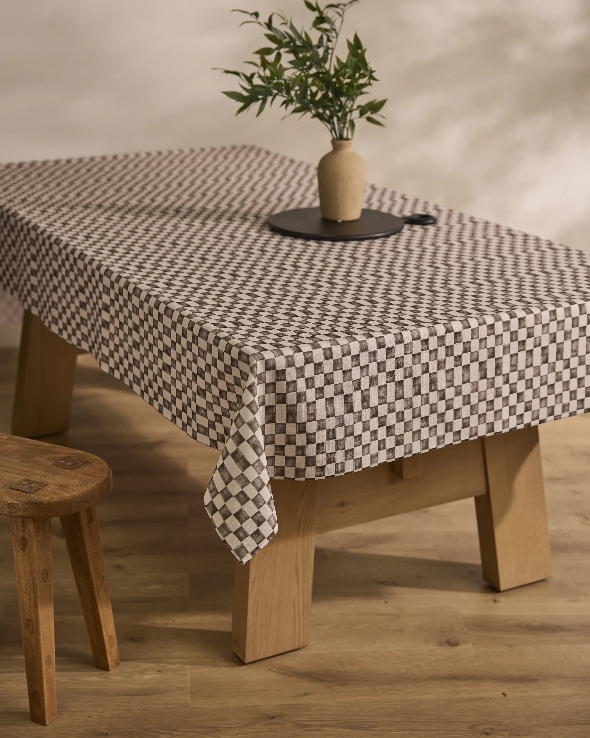 A Tavern wooden table by Geometry is topped with a stain-resistant black-and-white checkered tablecloth, a round black tray, and a ceramic vase with green leafy branches; a wooden stool sits beside it on a light wood floor.