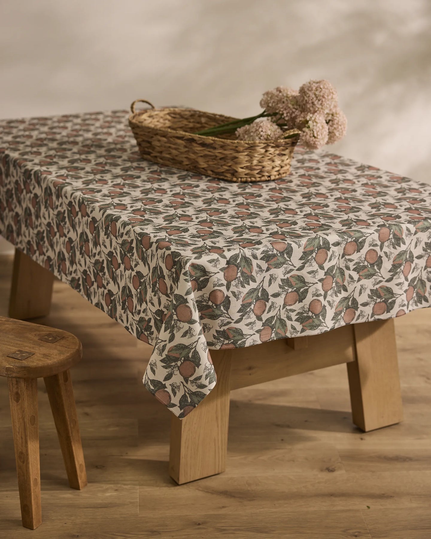 The Valencia tablecloth by Geometry, featuring pomegranates and leaves, covers a wooden table; on top sits a wicker basket with pale pink flowers, while a wooden stool stands beside the table on a wooden floor.