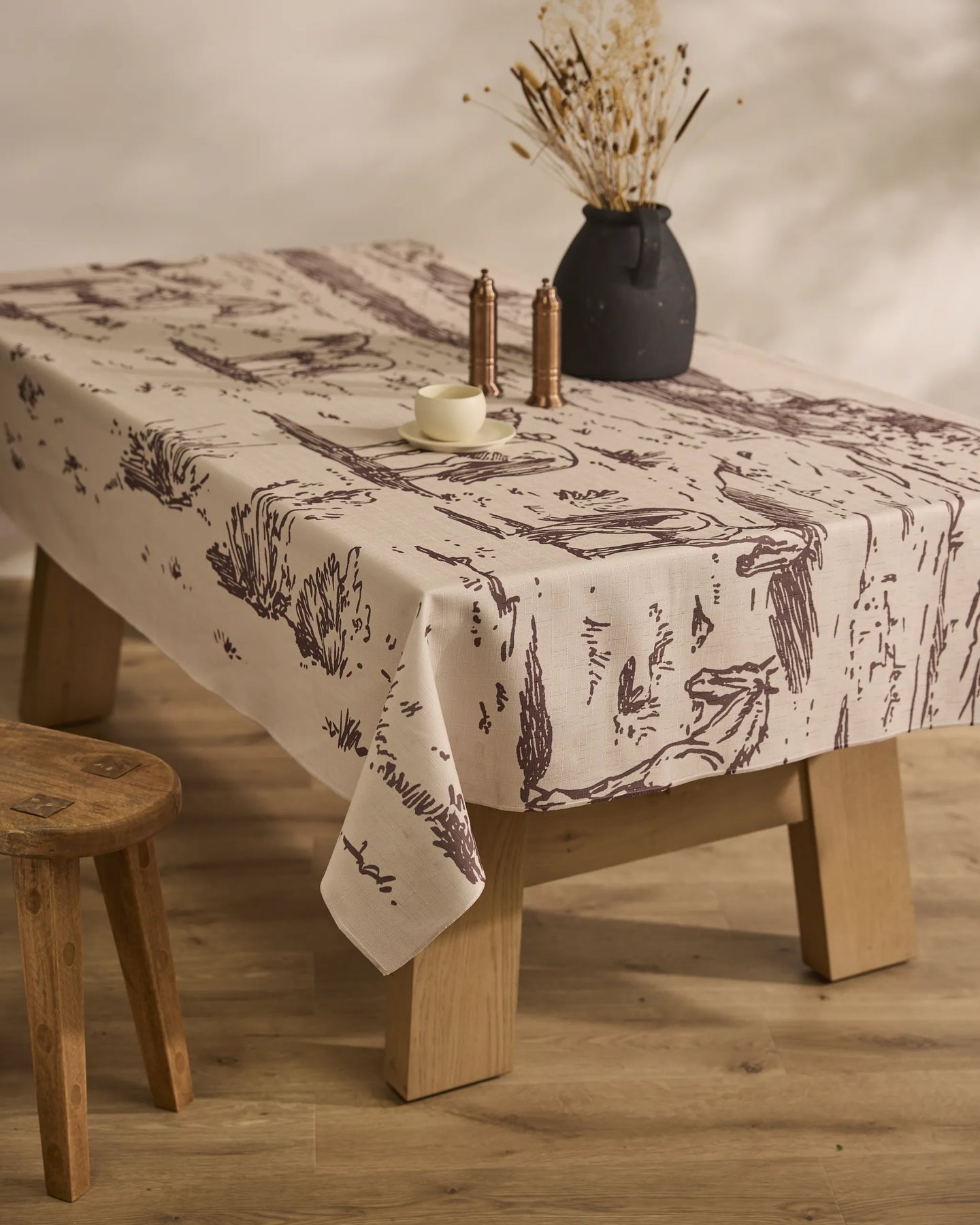 On a wooden table rests Geometrys Western Scene lightweight, stain-resistant tablecloth with nature designs. The setting includes a black dried-flower vase, two salt and pepper shakers, white cup and saucer, and a wooden stool on wood flooring.