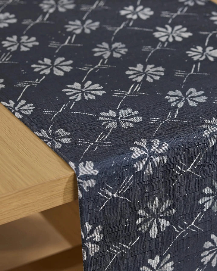 The Bonnie Indigo table runner by Geometry, featuring a stain-resistant dark blue fabric with a white floral pattern, is draped over the corner of a light wood table.