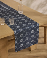 The Geometry Bonnie Indigo table runner, featuring a white floral pattern on dark blue, is stain-resistant and set on a wooden dining table with two clear glasses and a glass bottle. The floor is light wood.