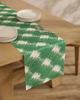 A light wood dining table features Geometrys Hairy Check Green stain-resistant runner, four stacked white plates, and two forks on top, set on a wooden floor.