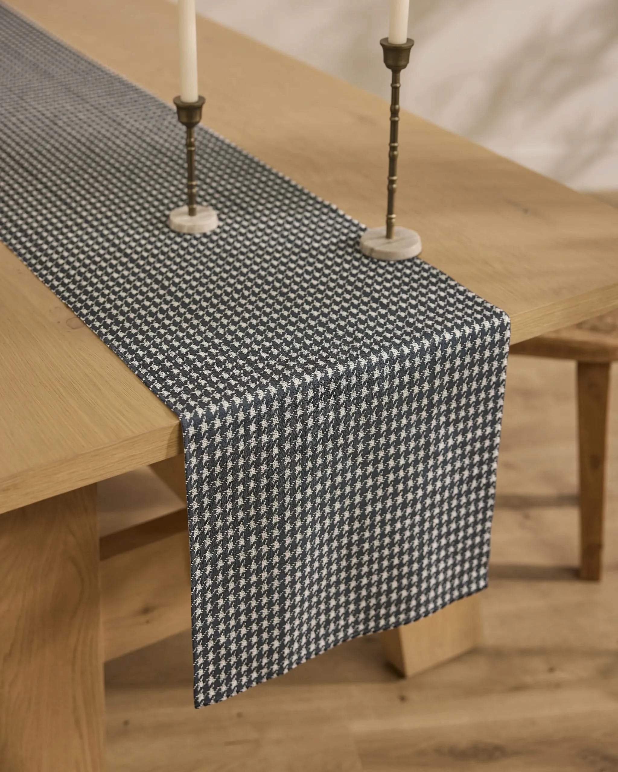 A wooden dining table is topped with the Geometry Houndstooth Black stain-resistant table runner, accented by two brass candlesticks with white candles, all placed on a light wood floor.