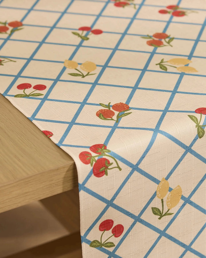 A close-up of a wooden table with Geometrys Italian Summer Grid table runner, featuring a blue grid and small red and yellow fruit with green leaves. Made from recycled materials, the runner drapes elegantly over the edge.