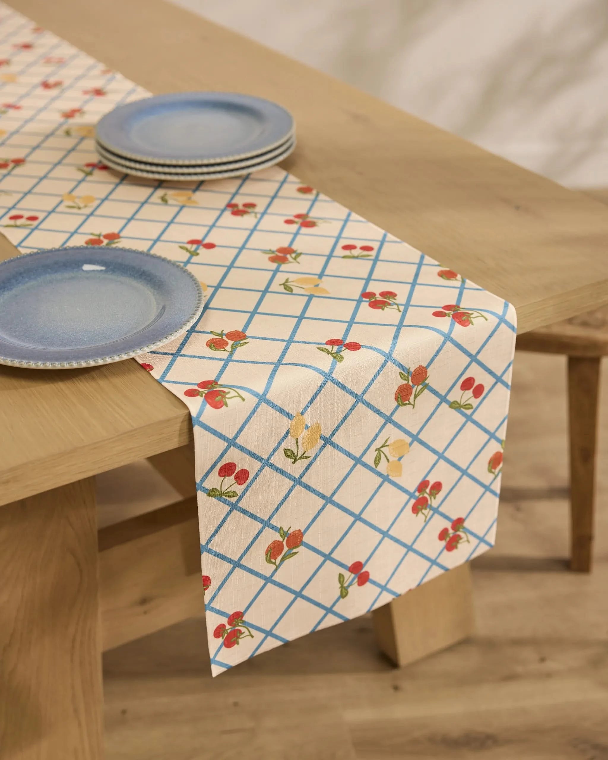 A wooden dining table features Geometrys Italian Summer Grid cream table runner, stain-resistant with blue grid and floral patterns. Blue ceramic plates are stacked at each setting.