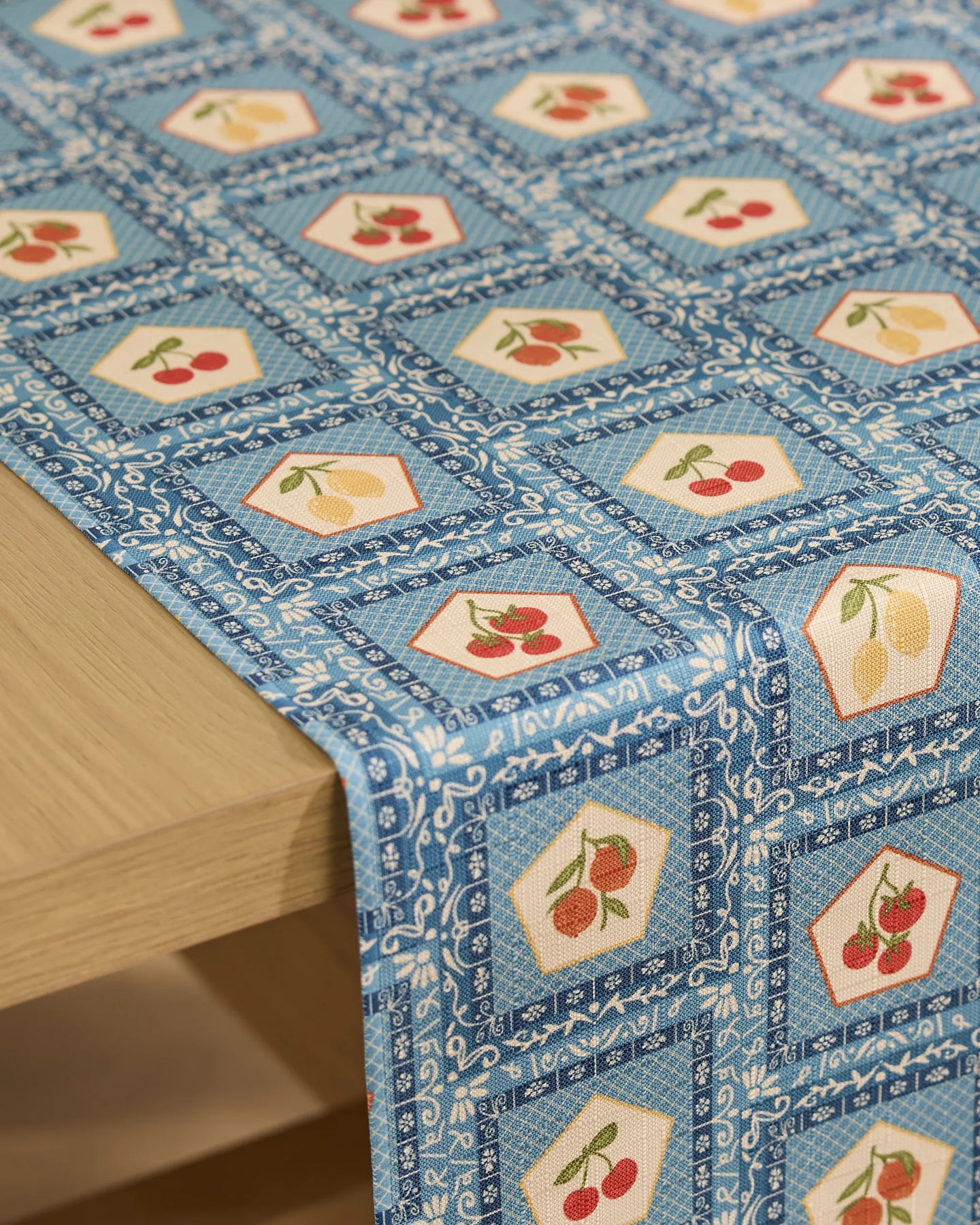 The Italian Tiles Table Runner by Geometry, featuring blue and white hexagonal patterns with cherries and lemons, drapes elegantly over a light wood table. This stain-resistant runner adds a fresh, modern touch to your dining decor.