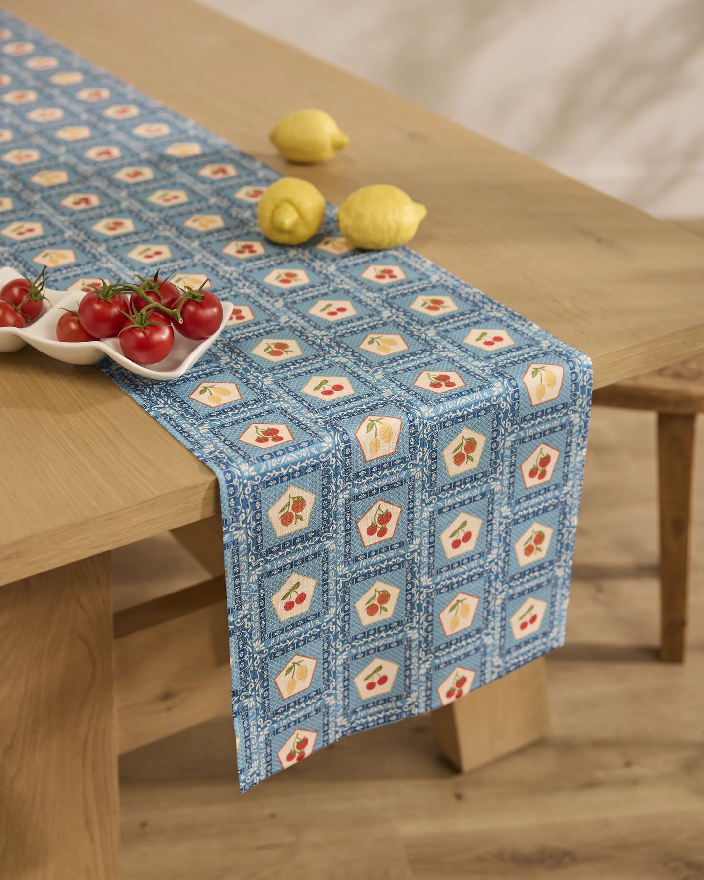 A wooden table with a Geometry Italian Tiles table runner, featuring blue tile designs, holds a ceramic dish of tomatoes and two lemons. The scene is set in a bright room with a neutral background.