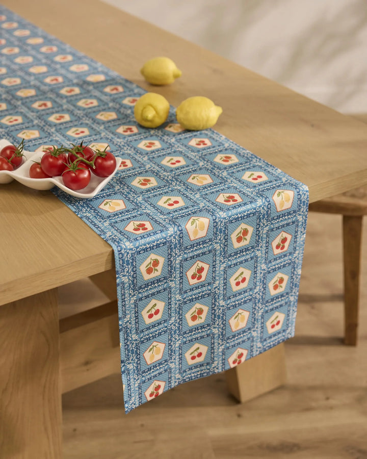 A wooden table with a Geometry Italian Tiles table runner, featuring blue tile designs, holds a ceramic dish of tomatoes and two lemons. The scene is set in a bright room with a neutral background.