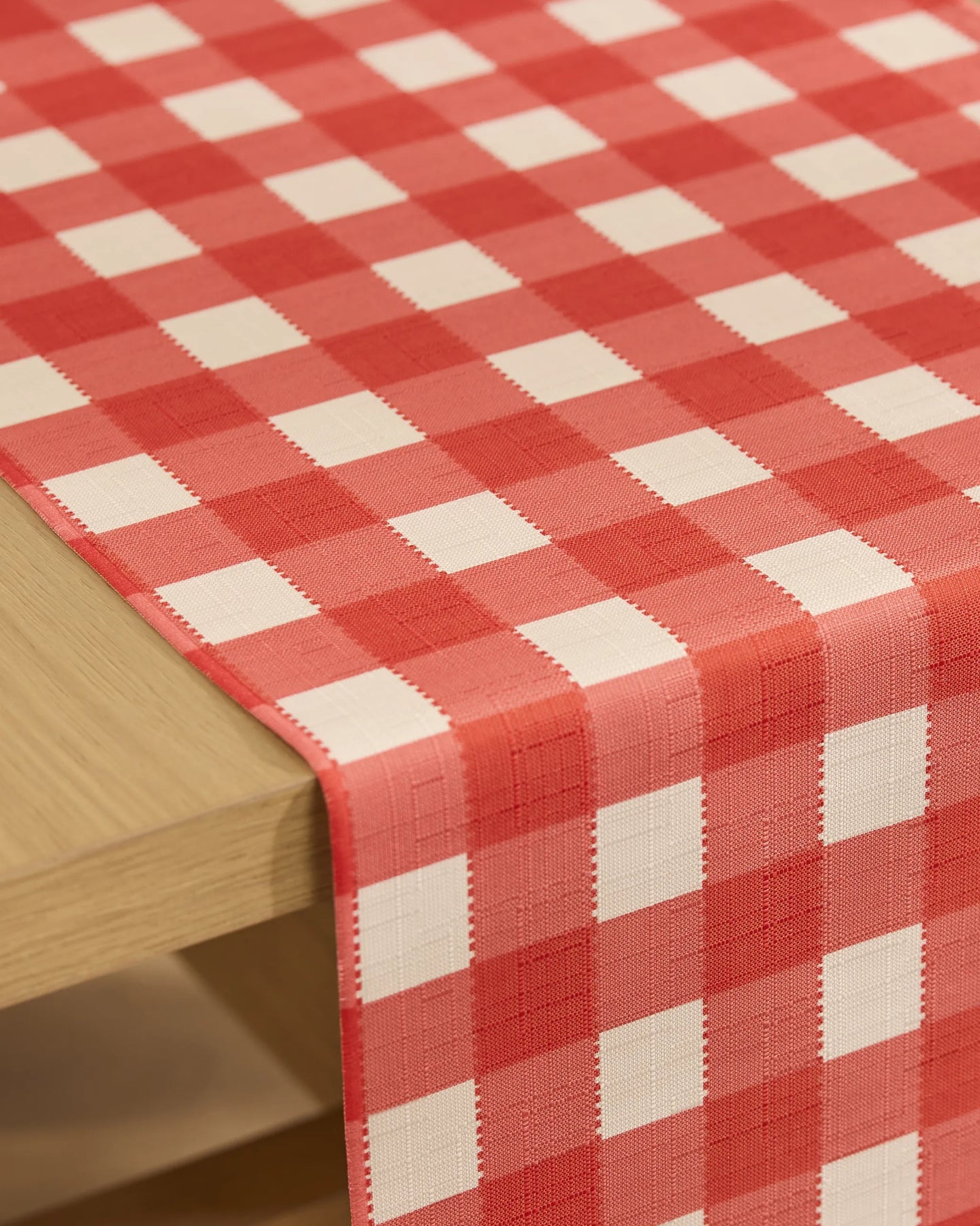A close-up of a wooden table with the Geometry Ripe Tomato Gingham stain-resistant table runner, featuring a classic red and white checkered pattern—perfect for everyday use.
