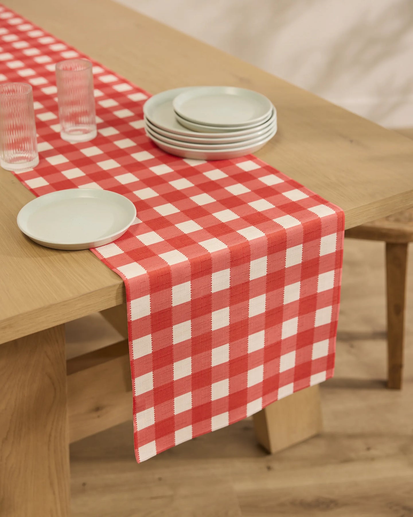 A wooden table set with Geometry’s Ripe Tomato Gingham stain-resistant table runner, three clear glasses, and a stack of white plates creates a cozy, casual dining atmosphere.