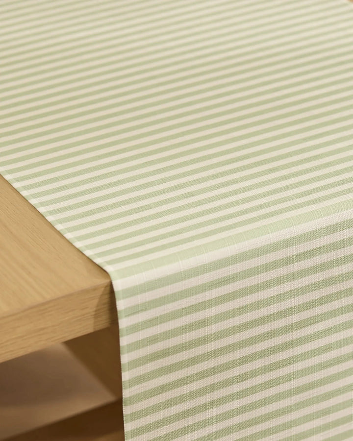 The Geometry Seagrass table runner, featuring light green and white stripes and stain-resistant fabric, is draped over the edge of a light wooden table.