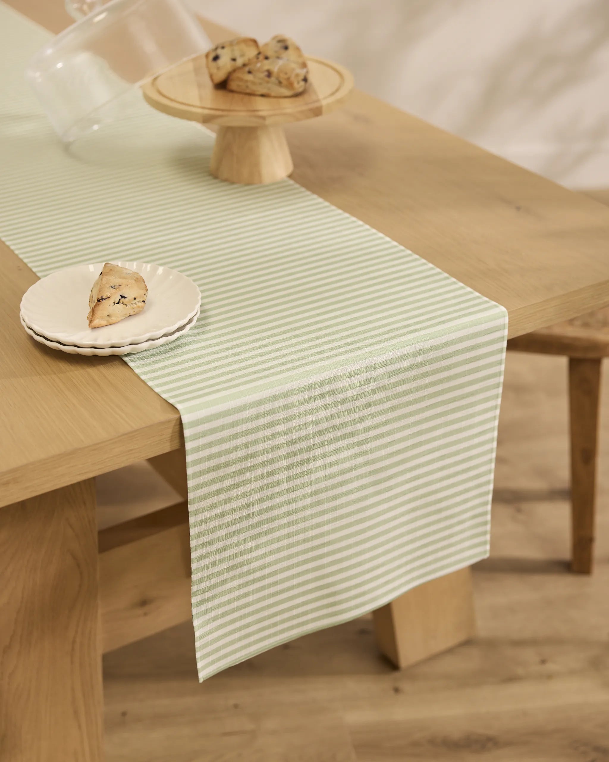 A light wood table features Geometrys Seagrass pale green and white striped stain-resistant runner. On top, a plate and platter each hold a scone, creating a warm, minimalistic, and inviting scene.