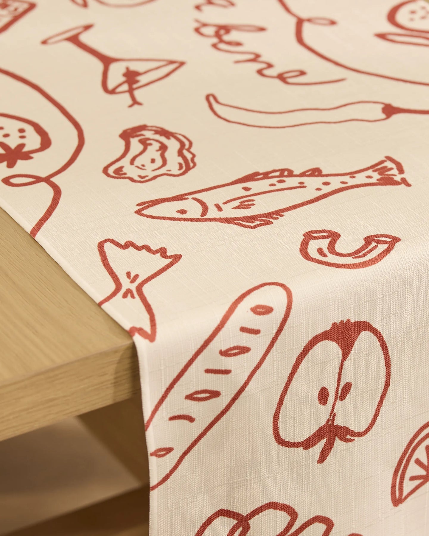 The Summer Air table runner by Geometry is beige, stain-resistant, and features simple red hand-drawn food illustrations, draped over the edge of a wooden table.