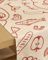 The Summer Air table runner by Geometry is beige, stain-resistant, and features simple red hand-drawn food illustrations, draped over the edge of a wooden table.