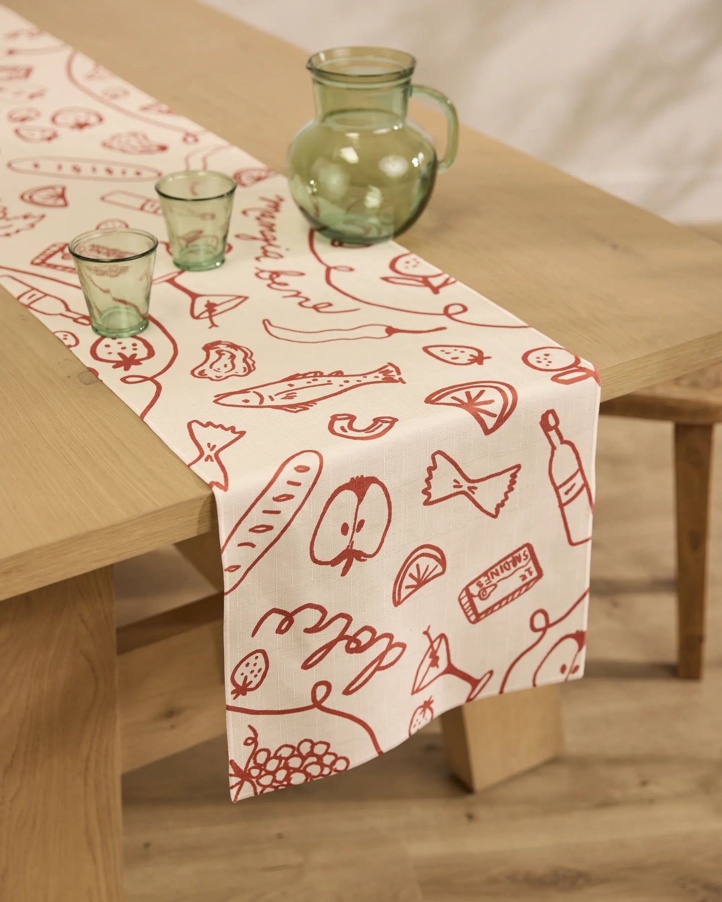 The Summer Air table runner by Geometry, featuring beige stain-resistant fabric with red food-themed illustrations, is laid on a wooden table set with green glassware in a softly lit room with wooden floors.