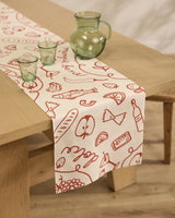 The Summer Air table runner by Geometry, featuring beige stain-resistant fabric with red food-themed illustrations, is laid on a wooden table set with green glassware in a softly lit room with wooden floors.