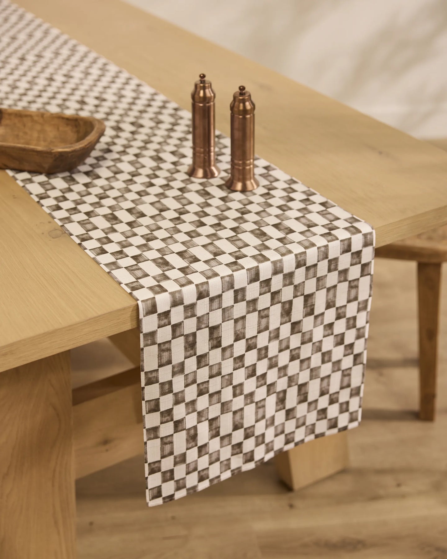 A Geometry Tavern dining table, topped with a black and white Geometry Table Runner, two copper salt and pepper shakers, and a wooden bowl, stands on a light wood floor.
