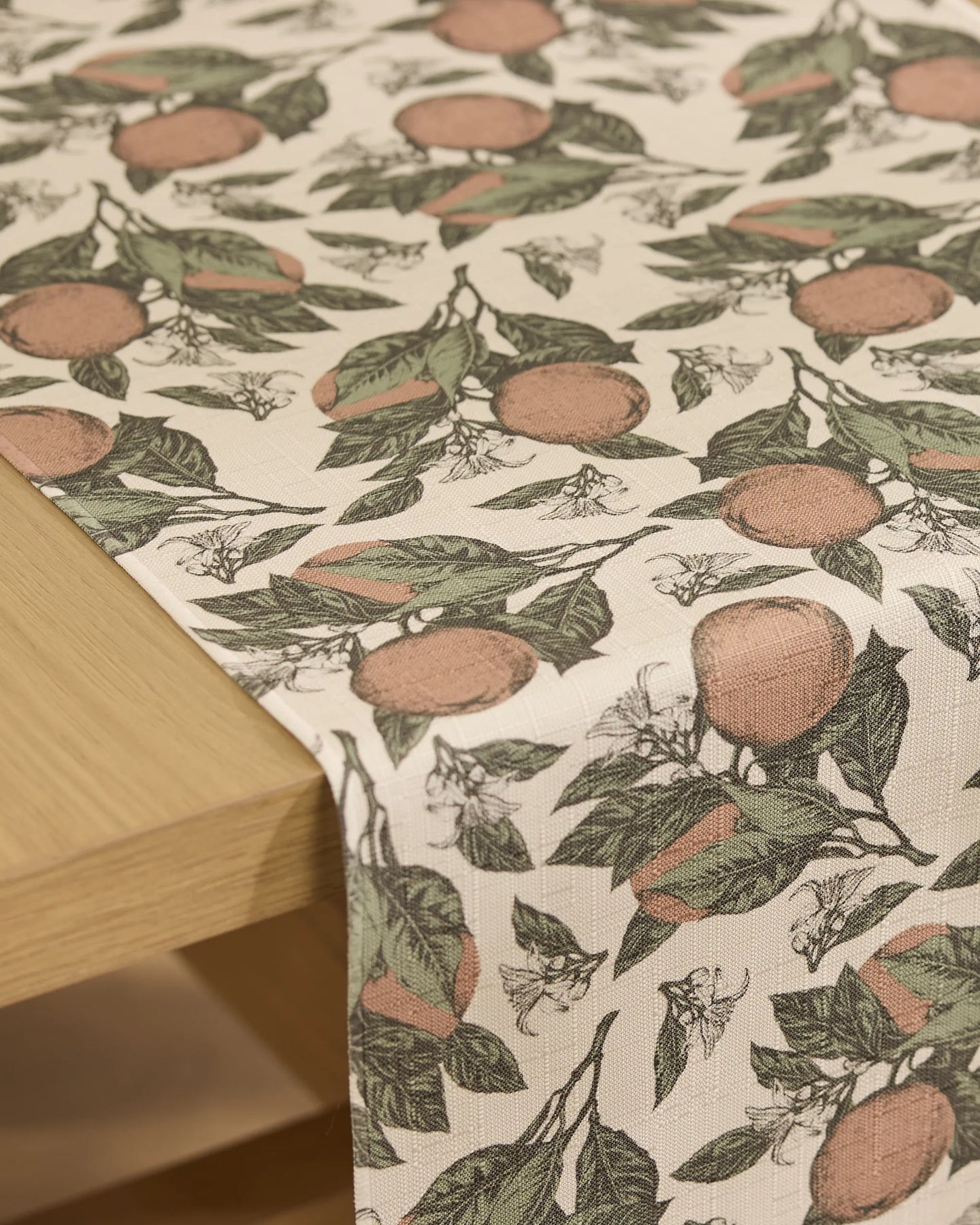 A close-up of the Valencia table runner by Geometry, featuring a stain-resistant light beige fabric with illustrated oranges, green leaves, and white blossoms, draped over a wooden table edge.