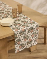 The Geometry Valencia stain-resistant table runner features an orange fruit and green leaf pattern. Two stacked white bowls and two brown glasses sit on it atop a wooden table with a light-colored wooden floor.