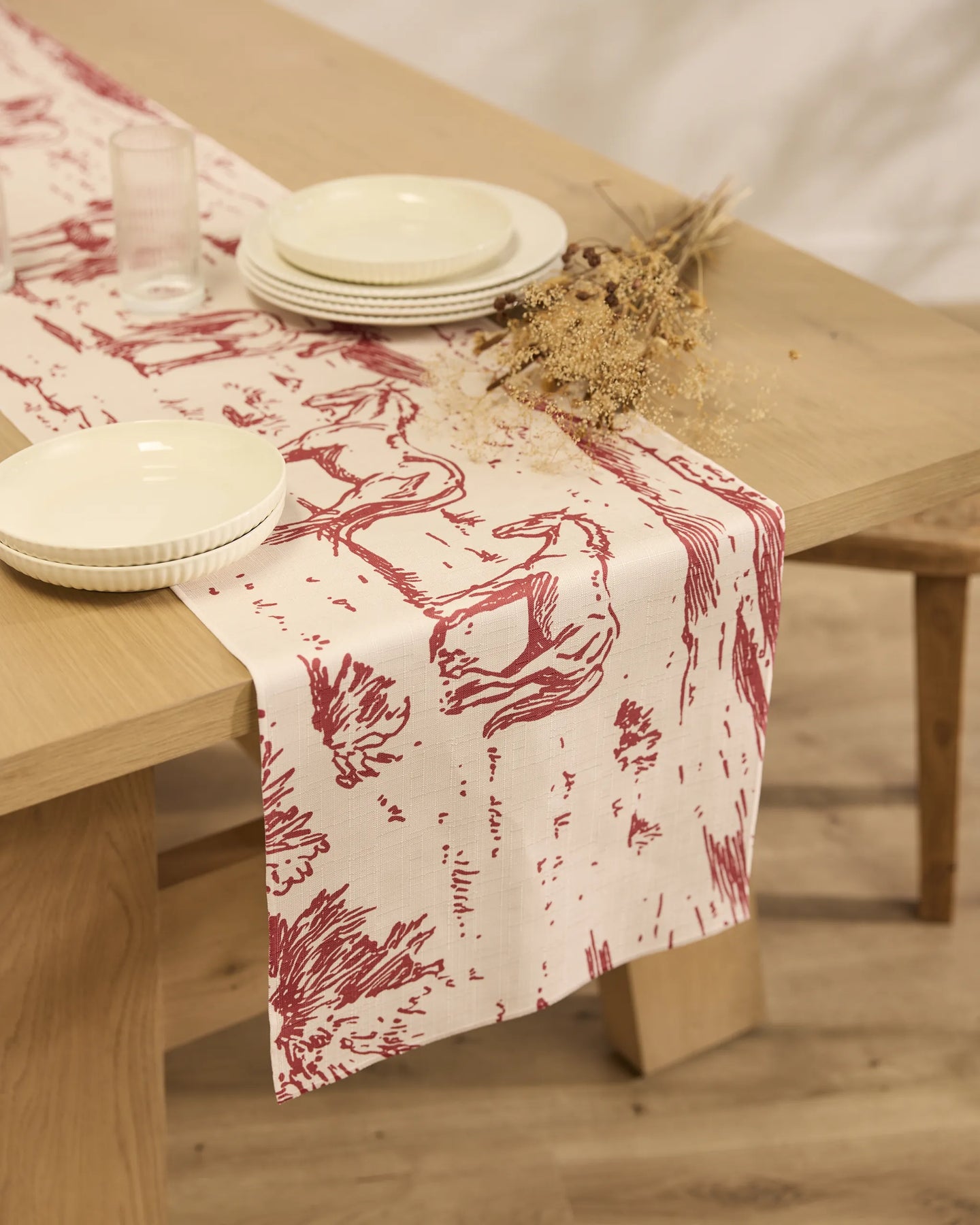 A wooden dining table features Geometrys Western Scene red and white patterned, stain-resistant table runner, neatly stacked cream-colored plates, a glass, and a small dried floral arrangement on top.