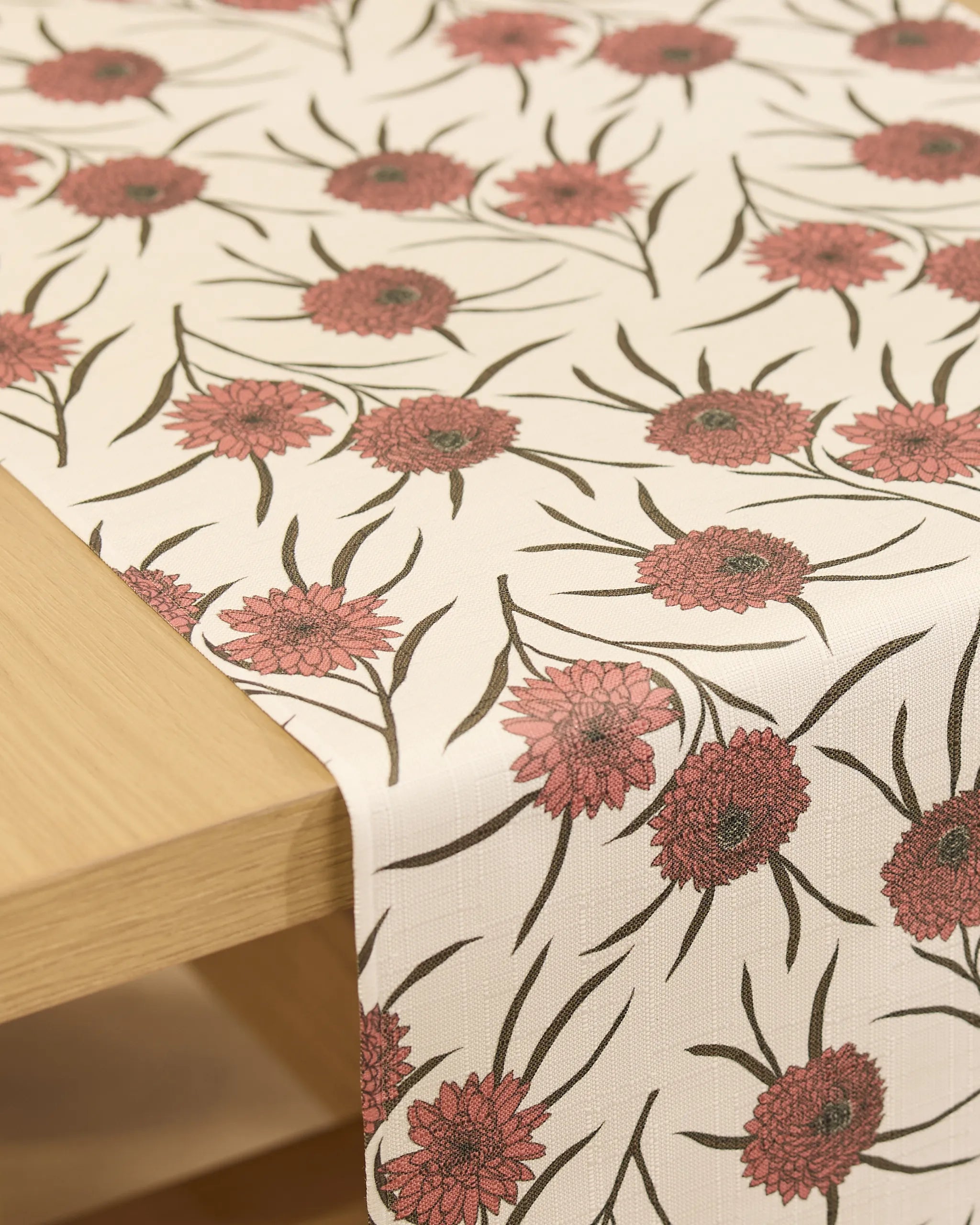 The Geometry Wild Dahlia table runner, cream-colored and stain-resistant with pinkish-red floral and green leaf patterns, is draped over a light wooden table.