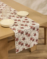 A light wooden table features a cream stain-resistant Wild Dahlia table runner by Geometry with red and green flowers. On the runner are two cream plates and four clear glasses, all set atop a light wood floor.