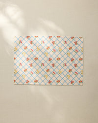 The Italian Summer Grid by Geometry, a rectangular sheet with a blue grid and small floral pattern, rests on a beige textured surface, its recycled placemat style highlighted by soft sunlight and gentle shadows.