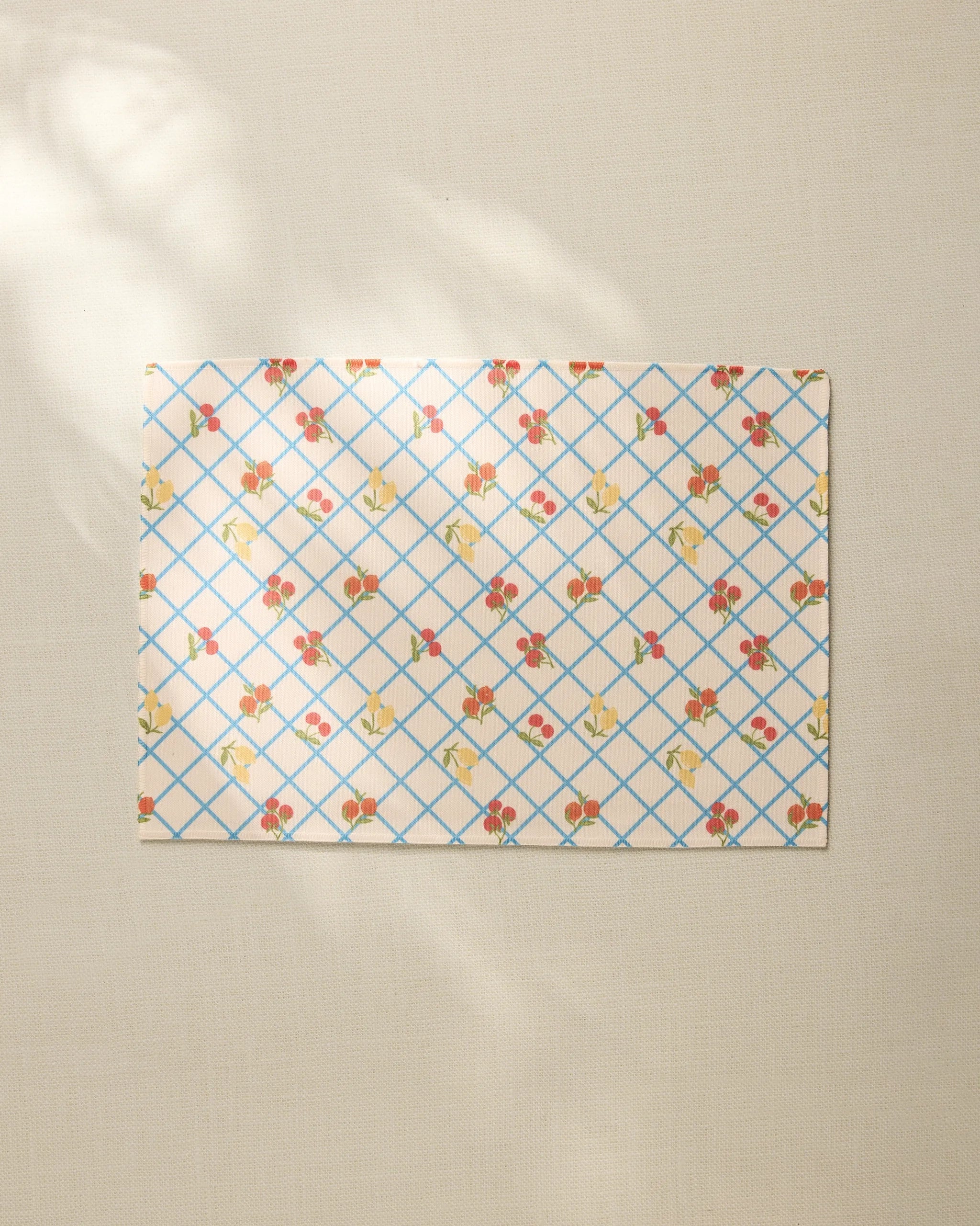 The Italian Summer Grid by Geometry, a rectangular sheet with a blue grid and small floral pattern, rests on a beige textured surface, its recycled placemat style highlighted by soft sunlight and gentle shadows.