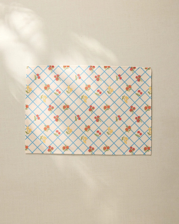 The Italian Summer Grid by Geometry, a rectangular sheet with a blue grid and small floral pattern, rests on a beige textured surface, its recycled placemat style highlighted by soft sunlight and gentle shadows.