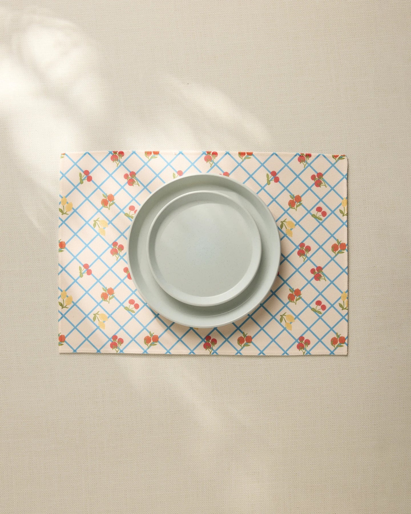 A plain light gray plate sits on the Geometry Italian Summer Grid Placemat, featuring a blue grid and small red and yellow floral pattern, atop a neutral tablecloth.