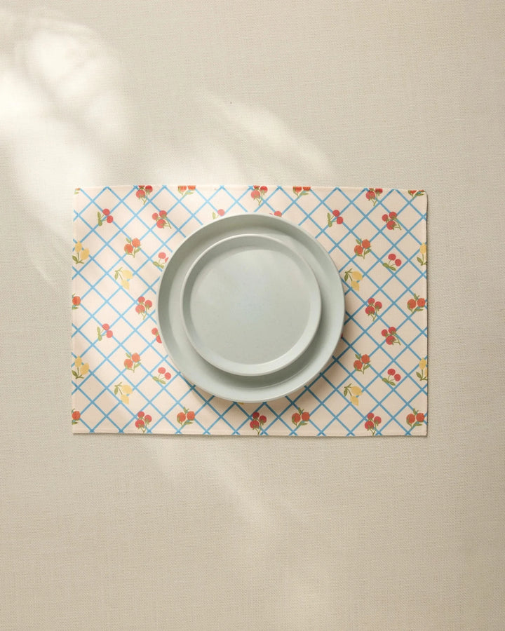 A plain light gray plate sits on the Geometry Italian Summer Grid Placemat, featuring a blue grid and small red and yellow floral pattern, atop a neutral tablecloth.