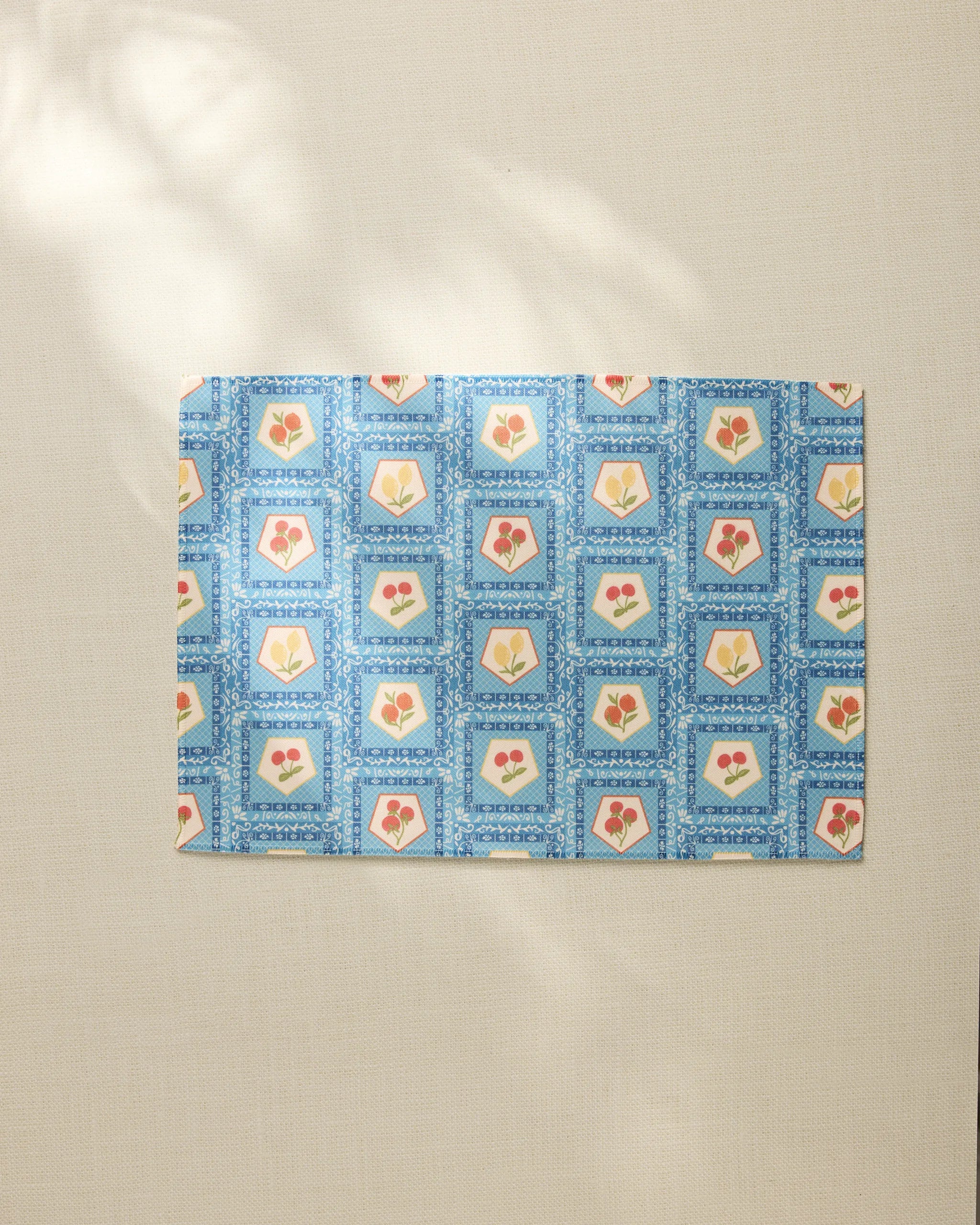 The Geometry Italian Tiles placemat features a blue geometric border and square patterns with red and yellow floral or heart motifs, arranged on a light beige surface.