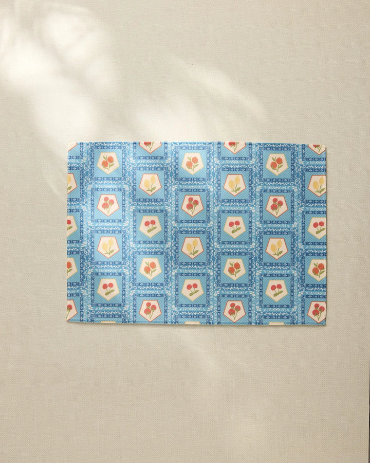 The Geometry Italian Tiles placemat features a blue geometric border and square patterns with red and yellow floral or heart motifs, arranged on a light beige surface.