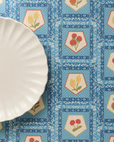 A white scalloped-edge plate rests on a Geometry Italian Tiles blue patterned tablecloth featuring hexagonal motifs with red and yellow flowers, paired with matching recycled placemats.