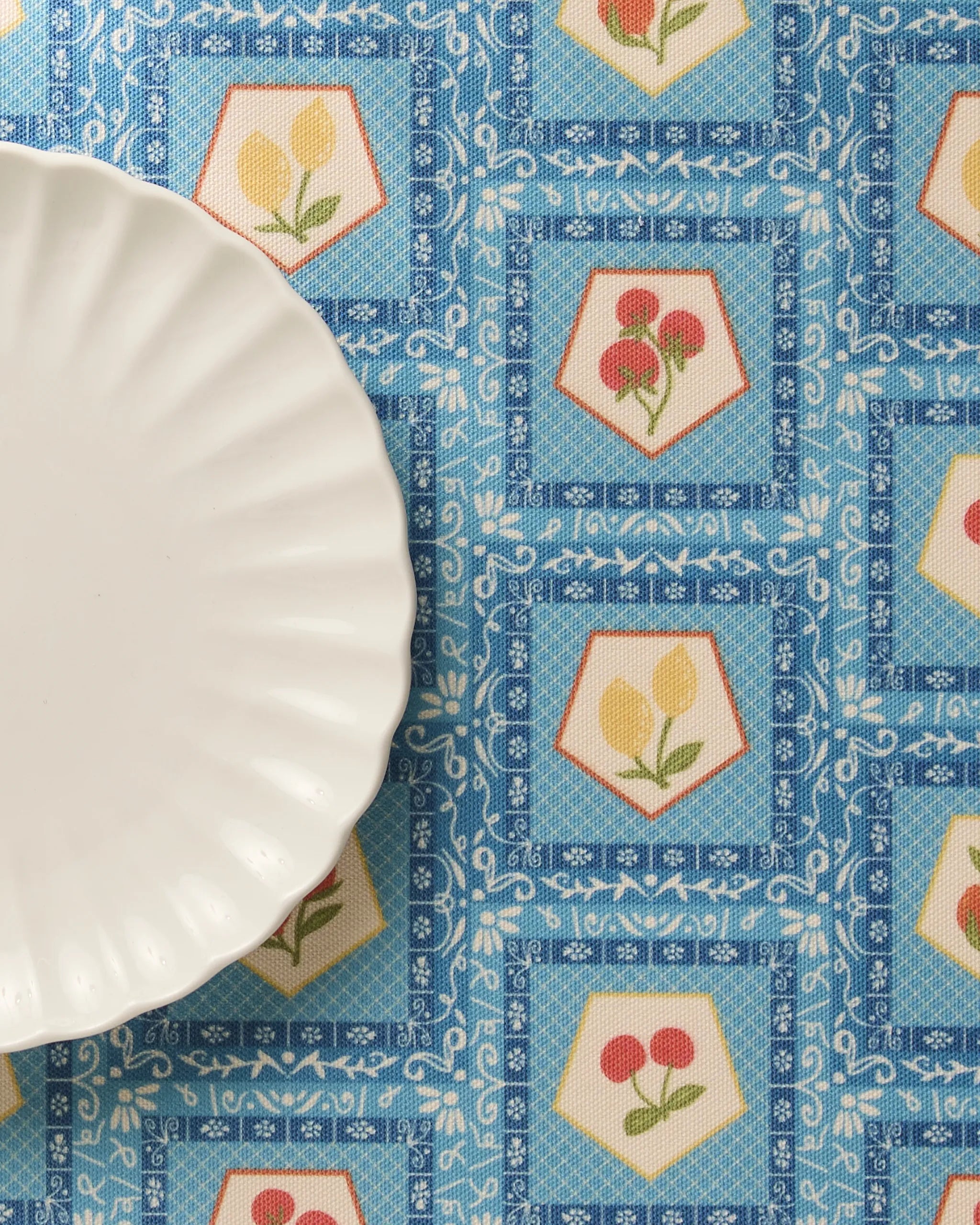 A white scalloped-edge plate rests on a Geometry Italian Tiles blue patterned tablecloth featuring hexagonal motifs with red and yellow flowers, paired with matching recycled placemats.