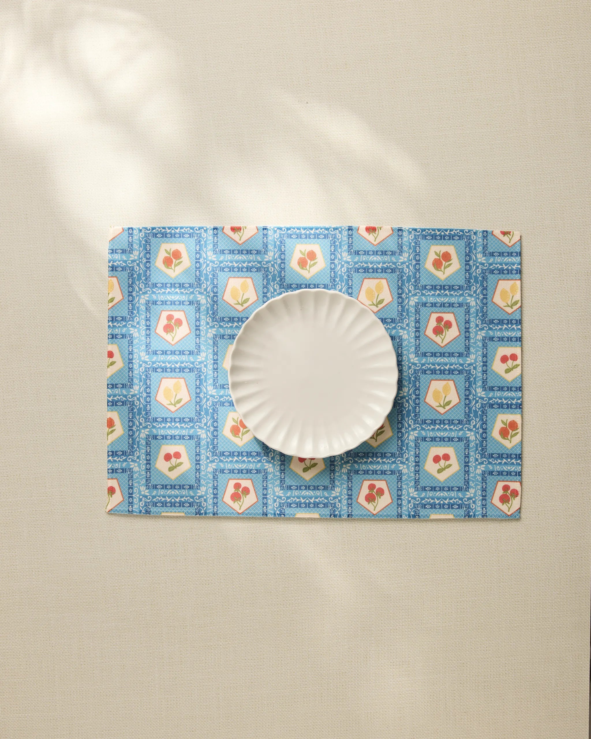 The Geometry Italian Tiles fluted plate rests on a blue floral placemat made from recycled materials atop a light tablecloth, with sunlight casting gentle shadows across the scene.
