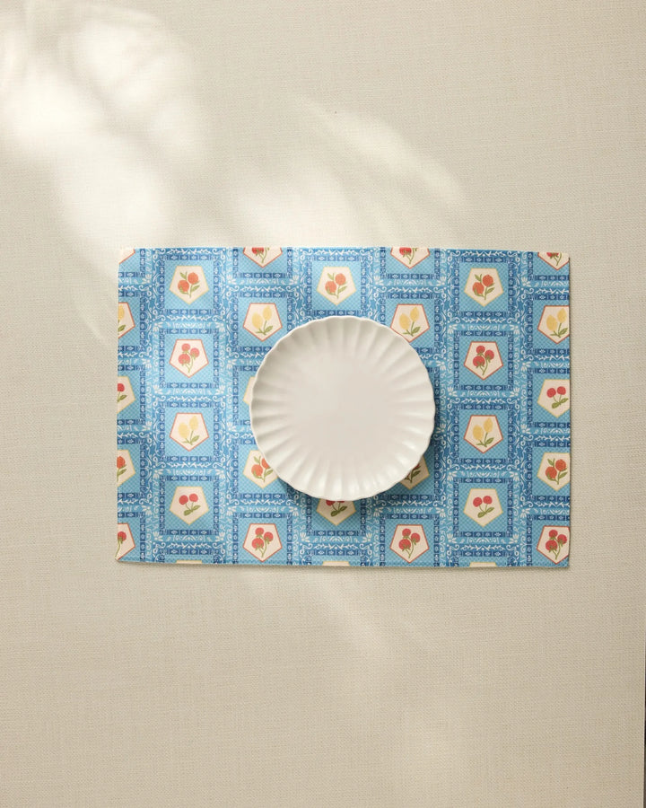 The Geometry Italian Tiles fluted plate rests on a blue floral placemat made from recycled materials atop a light tablecloth, with sunlight casting gentle shadows across the scene.