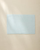 A Geometry Chambray Light Blue rectangular placemat is laid flat on a beige surface, illuminated softly by natural light from the left.