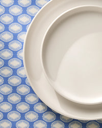 Two white ceramic plates, one large and one small, from Geometrys Lagoon line, are stacked on a blue tablecloth featuring a white sunburst pattern. The plates are placed in the upper right corner of the image.