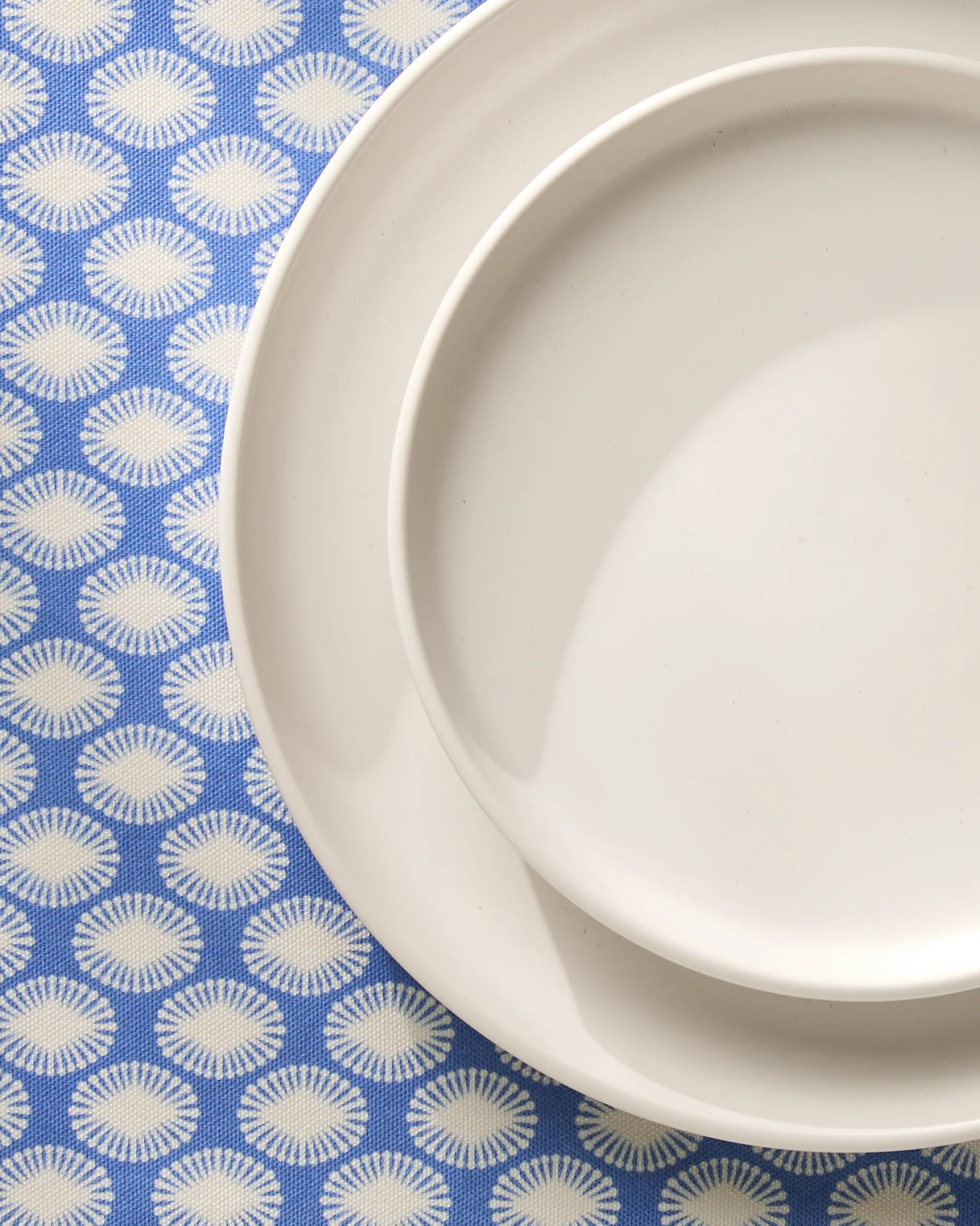 Two white ceramic plates, one large and one small, from Geometrys Lagoon line, are stacked on a blue tablecloth featuring a white sunburst pattern. The plates are placed in the upper right corner of the image.