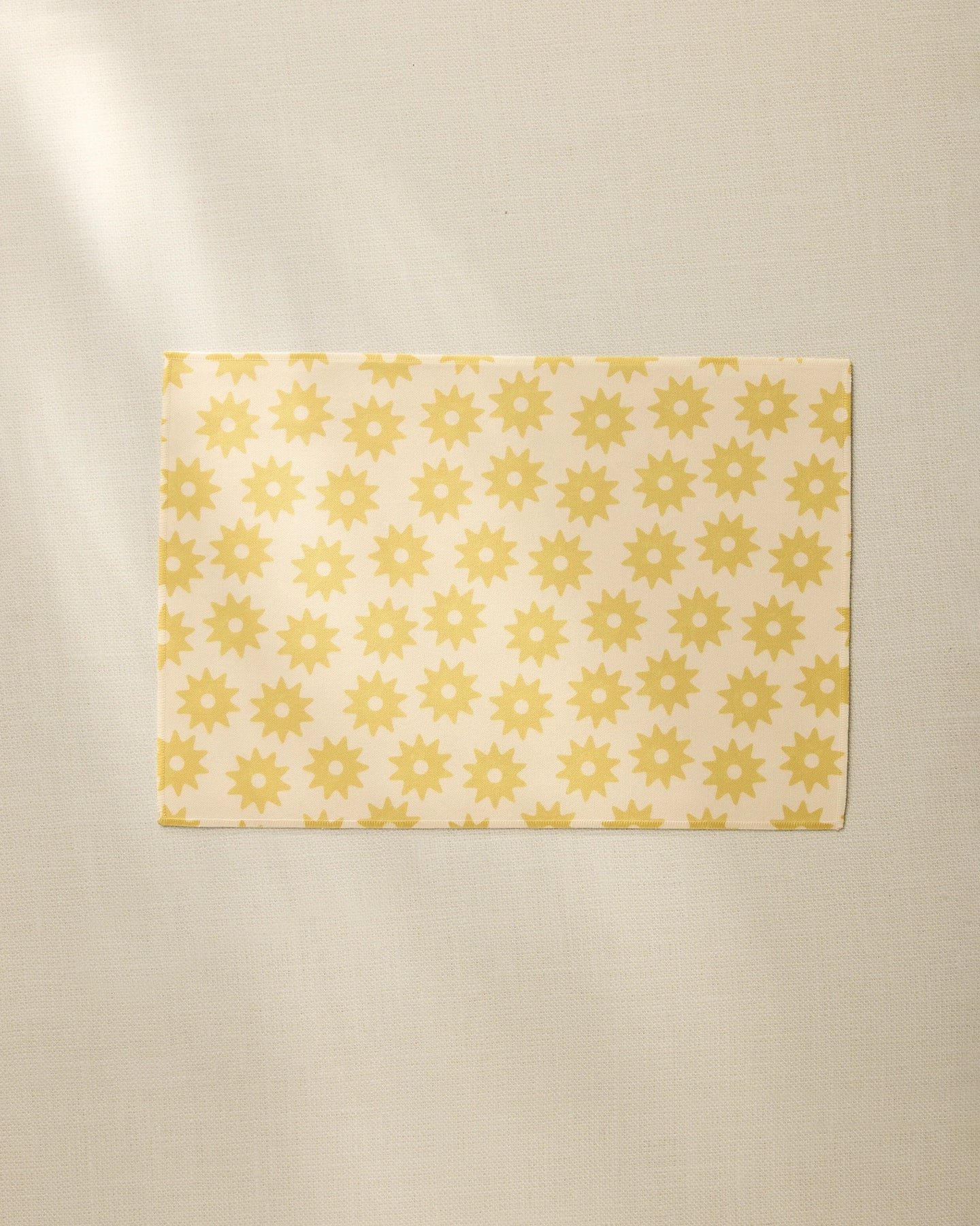 The Geometry Sunny Sun recycled placemat, with a repeating yellow sunburst pattern, rests on a light beige fabric surface in soft natural light.