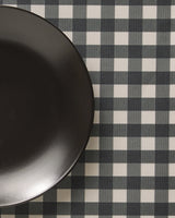 A black plate sits on the left side of a checkered tablecloth with a black and white Charcoal Gingham pattern from Geometry, featuring stylish, machine washable placemats for easy care.
