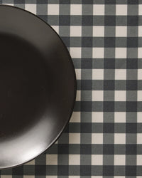 A black plate sits on the left side of a checkered tablecloth with a black and white Charcoal Gingham pattern from Geometry, featuring stylish, machine washable placemats for easy care.