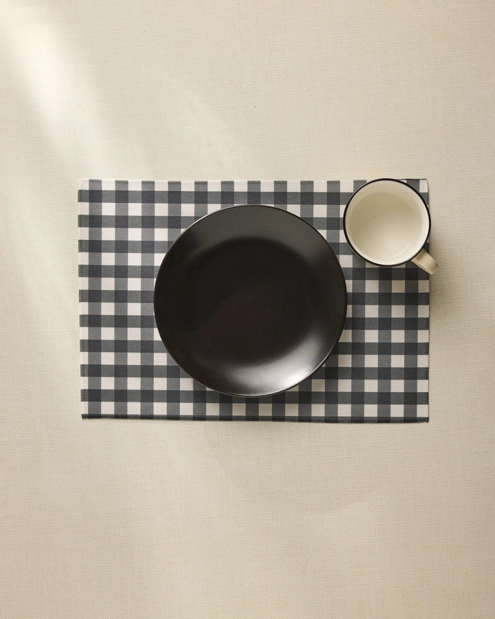 A black bowl and white mug rest on the Charcoal Gingham placemat by Geometry, neatly arranged atop a light-colored tablecloth.