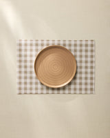 A round, light brown ceramic plate sits centered on the Geometry Hazel Gingham Placemat atop a cream-colored tablecloth.