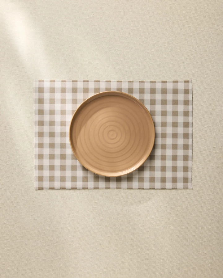 A round, light brown ceramic plate sits centered on the Geometry Hazel Gingham Placemat atop a cream-colored tablecloth.